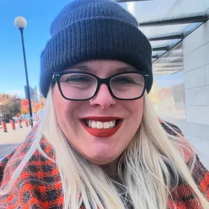 Kelsey Seguin, with glasses and long blonde hair, wears a blue knit hat and a red plaid coat. She smiles, outdoors on a city street.