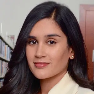Sarah Raza, with long dark hair, wears a light blazer and stands beside library bookshelves, looking at the camera.