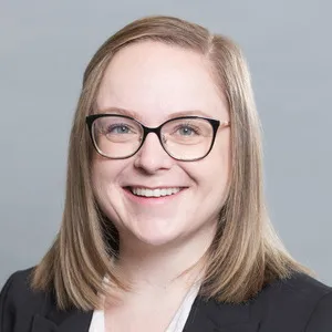 Brittany Finlay, with glasses and shoulder-length blonde hair, wears a black blazer and smiles at the camera against a plain background.