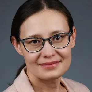 Alfiya Battalova, with dark hair pulled back and glasses, looks toward the camera with a smile, wearing a light blazer over a patterned top.