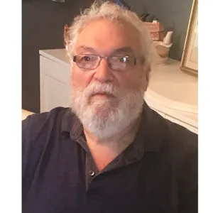 Sid Frankel with curly white hair, a full white beard, and glasses, looks at camera while seated indoors, wearing a dark polo shirt.