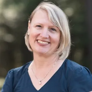 Tracy Smith-Carrier, with short blonde hair, smiles at the camera in an outdoor portrait, wearing a navy blouse and a small pendant necklace with trees softly blurred in the background.