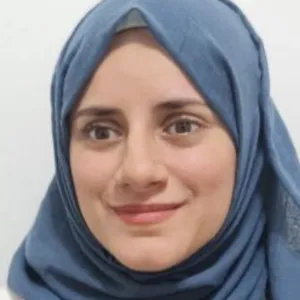 Aya Morad, wearing a blue hijab and a floral-patterned top, looks toward the camera with a smile against a plain background.