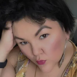 Kelly Johnson, with short dark hair, rests her head on her hand and looks directly at the camera, wearing long earrings and a patterned top.