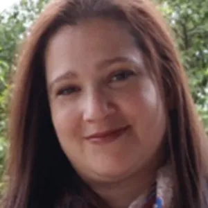 Lina Gharbiya, with long brown hair, smiles gently at the camera in an outdoor portrait, wearing a patterned scarf with green trees blurred in the background.
