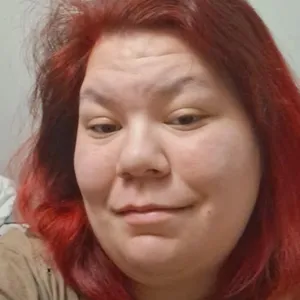 Skyler Wolf, with shoulder-length red hair, looks into the camera while taking a selfie indoors, wearing a tan shirt against a wall background.