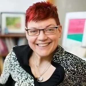 Nancy Hansen, with short red hair and glasses, smiles at the camera in a room with bookshelves in the background.