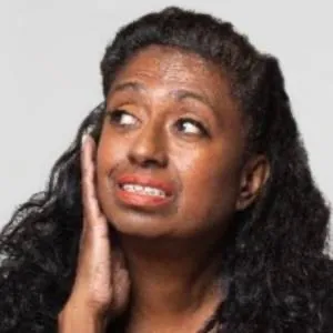 Doris Rajan, with long dark curly hair, rests her hand on her cheek and looks upward with a thoughtful expression.