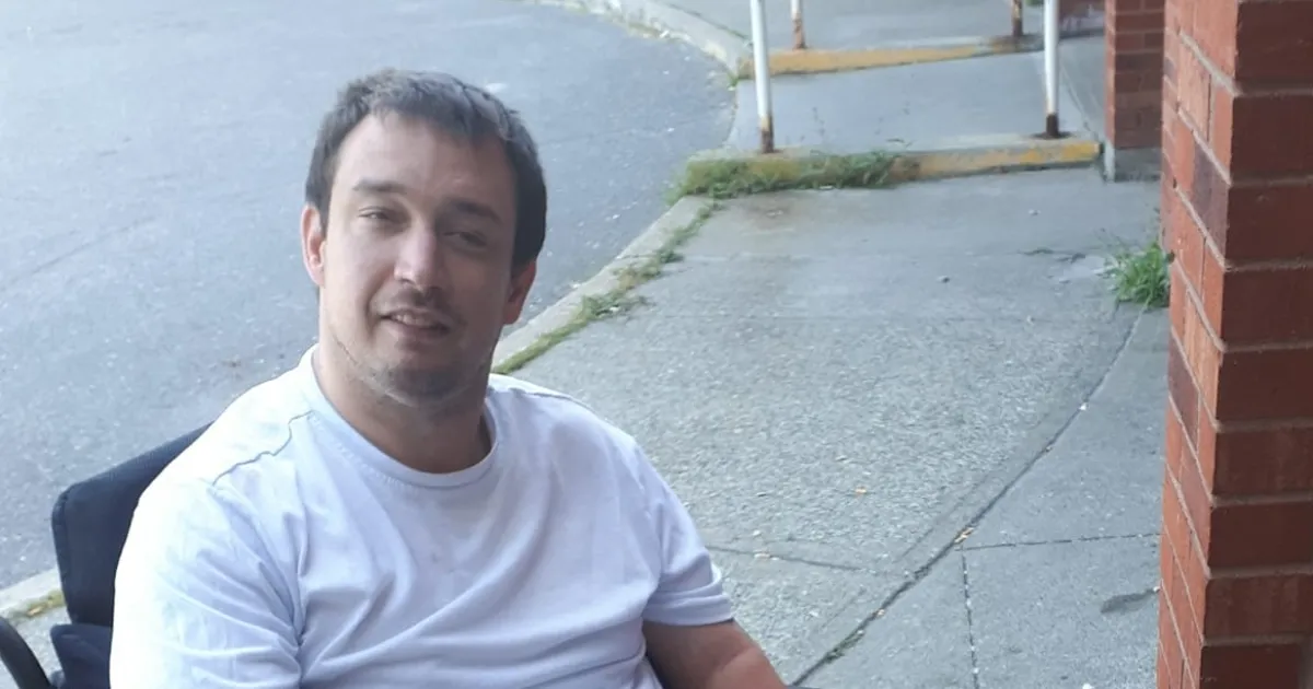 White man seated in wheelchair with short dark brown hair, a mustache, and wearing a white tshirt.
