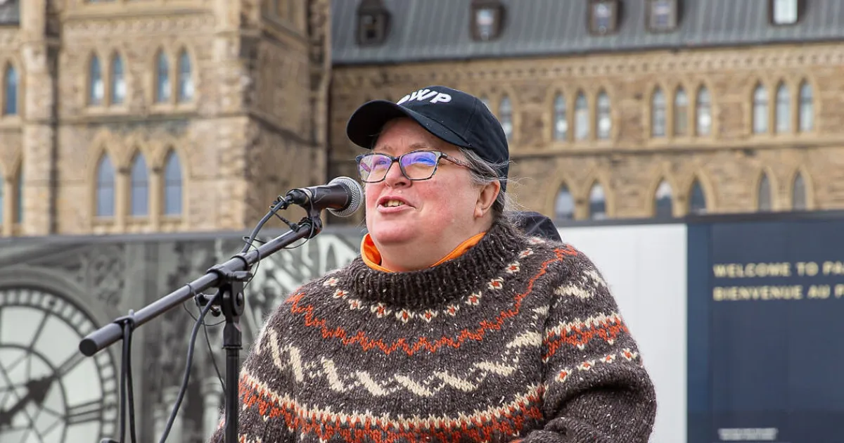 White woman wearing black baseball cap and glasses is seated in a wheelchair. She is wearing a brown sweater and is speaking into a mike.