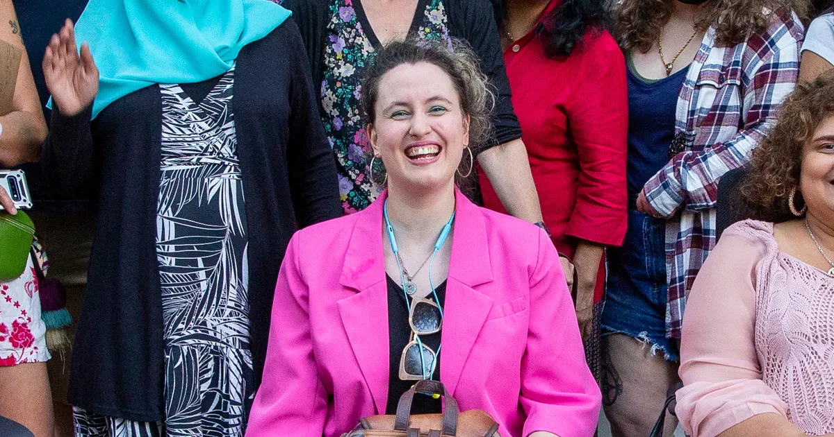 White woman with curly short hair seated in wheelchair. She is wearing a pink blazer and black top.